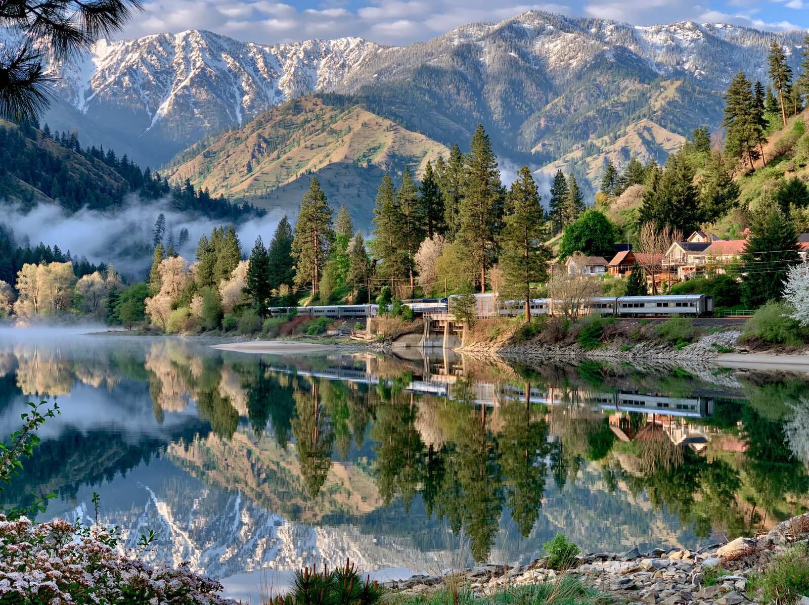 A train running along the shore of a mountain lake, the forest and snowy peaks mirrored in the still water
